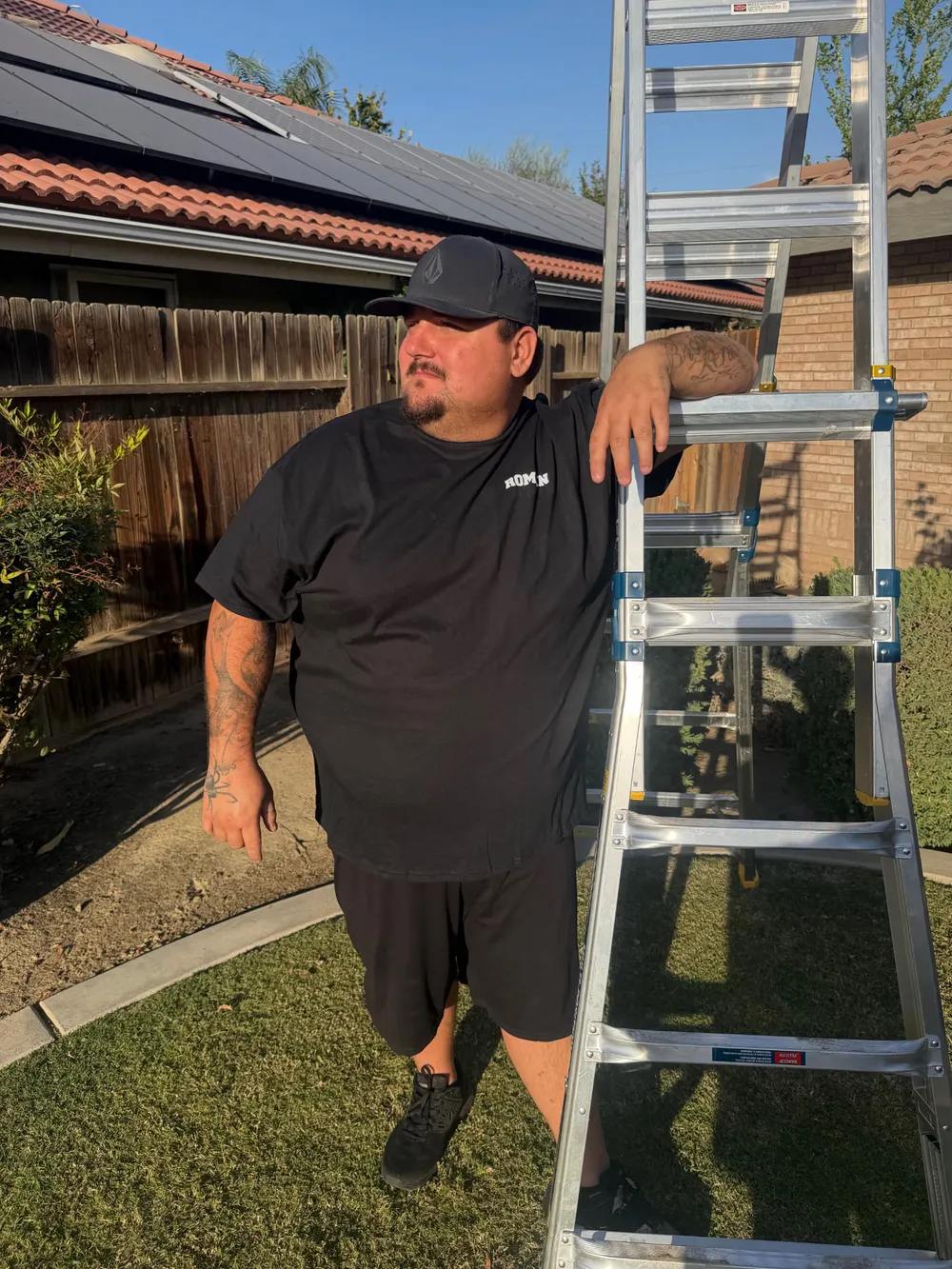 Roman of Suds and Studs leaning on a ladder in a backyard with rooftop solar panels