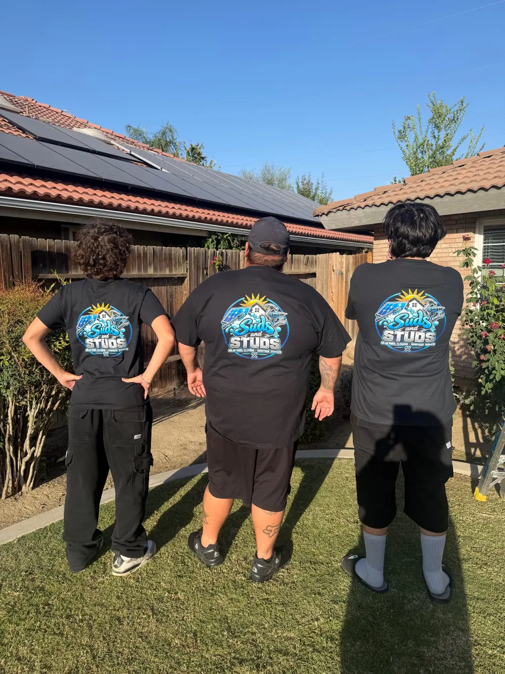Three Suds and Studs team members in logo shirts looking up at rooftop solar panels