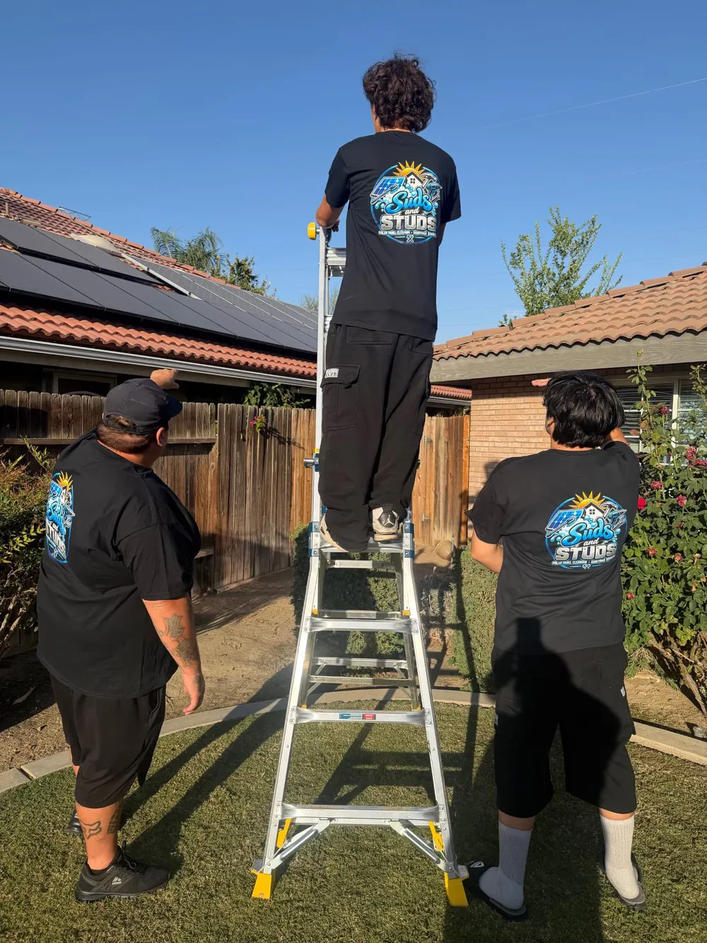 Suds and Studs crew member on a ladder inspecting a solar-paneled roof while two teammates guide